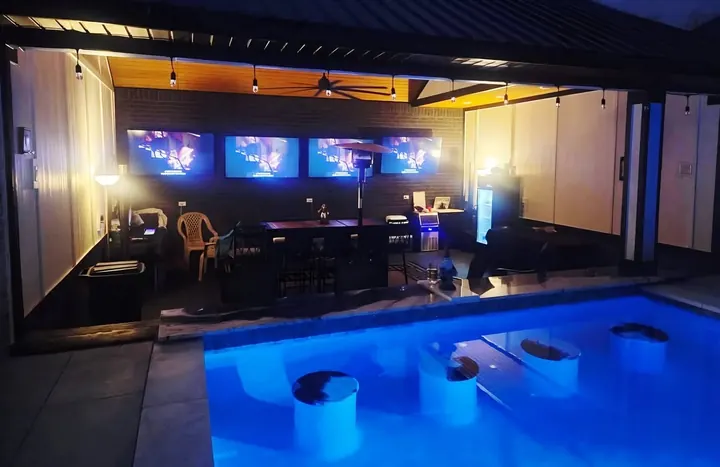 Outdoor bar lit at night with a row of TVs above a glowing blue pool