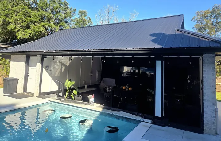 Pool house with a black metal roof and glass doors beside a pool with swim-up stools